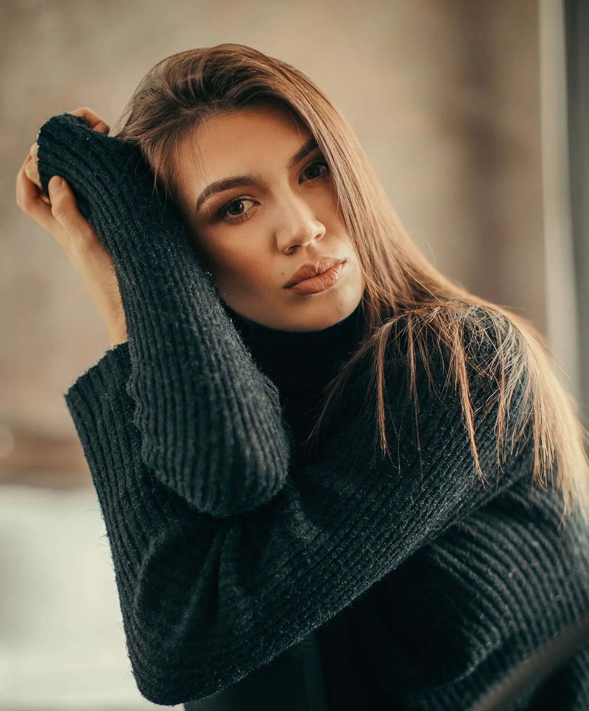 A serious-looking young woman with long, brown hair is posing while wearing a dark, ribbed turtleneck sweater. She is resting her chin on her hand, with her arms crossed and tucked into her sweater sleeves. Her gaze is directed at the camera with a confident expression. The background is a soft-focus, indoor setting with warm, natural light. - Blepharoplasty in Houston, TX