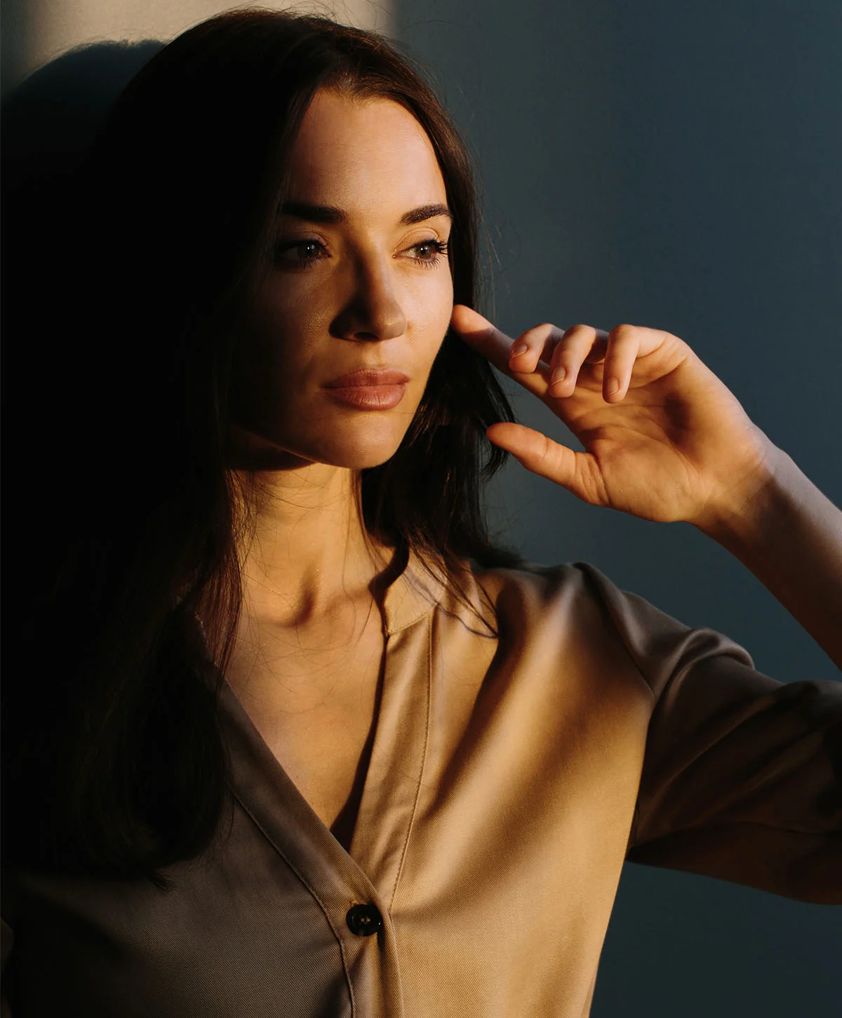 A horizontal shot of a young woman with long, dark hair, wearing a light gray collared shirt. She is standing with a pensive expression, looking to the side. There is a strong contrast of light and shadow, with a beam of sunlight hitting the right side of her face and hair. The background is a solid dark color. - Botox in Houston, TX