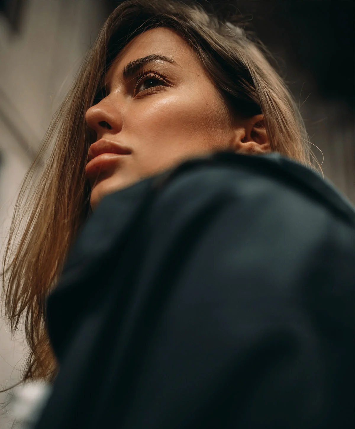 A low-angle shot of a beautiful young woman looking up and to the side, with a determined or pensive expression. She has long, light brown hair and is wearing a dark jacket. The background is blurred, suggesting an urban or modern setting, possibly indoors. The lighting highlights her face, focusing on her brow, eyes, and cheekbones. - Brow Lift in Houston, TX