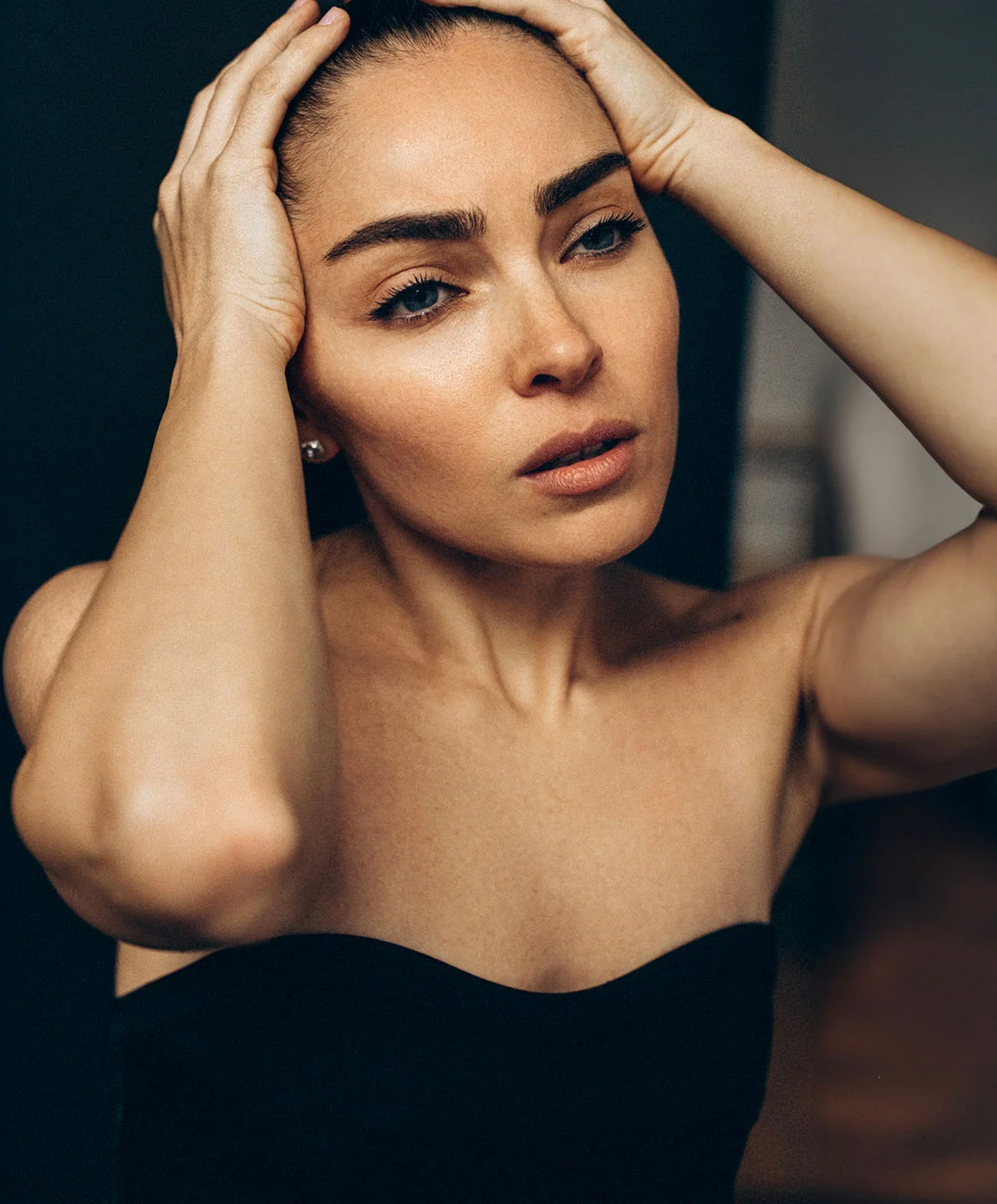 A young woman in a black strapless top holds her head in her hands, looking distressed, against a dark background. - Buccal Fat Removal in Houston, TX