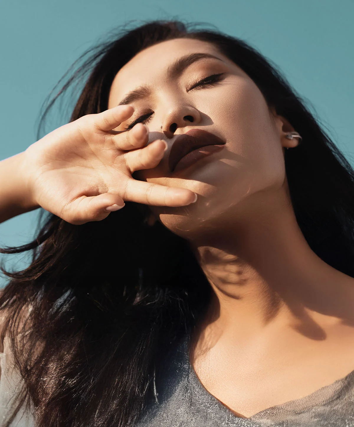 A striking, low-angle portrait of a woman with long dark hair, closing her eyes and looking up at the sky. She is wearing a light-colored top and holding one hand partially over her face, with sunlight and shadow accentuating her features. - Buccal Fat Removal Recovery in Houston, TX
