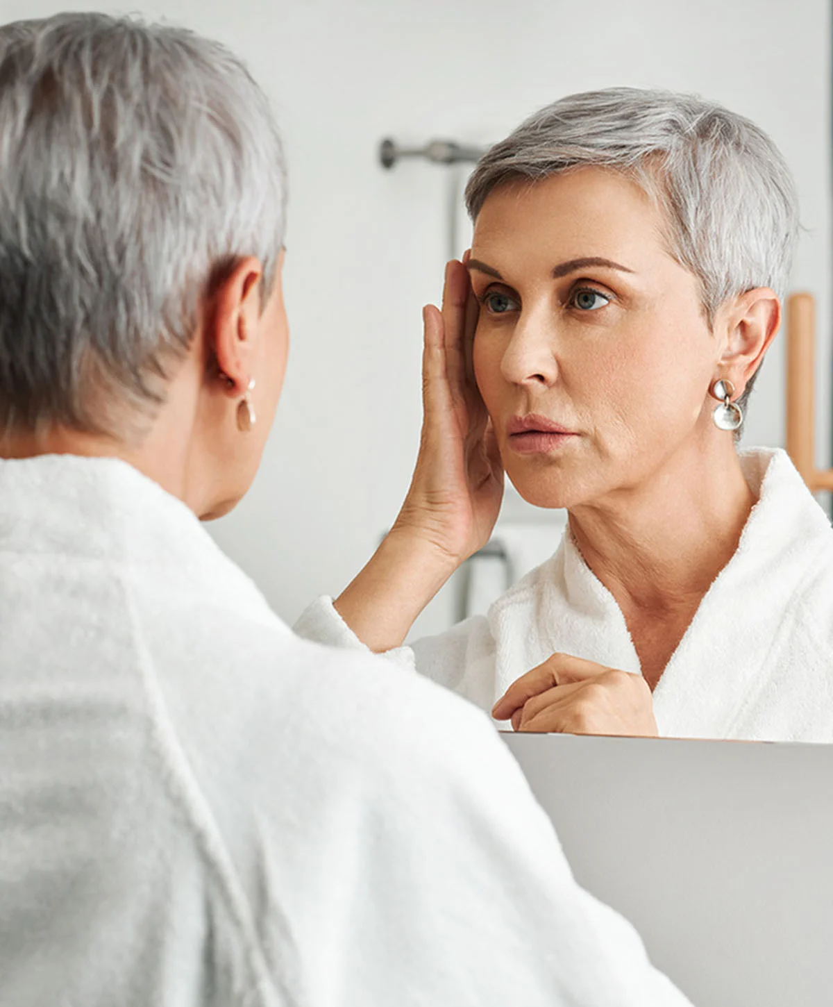 A woman with short, gray hair is looking at her reflection in a mirror. She is wearing a white bathrobe. She is touching her face, and her expression appears somewhat concerned or pensive. The lighting in the bathroom is bright and neutral. The background is a light gray/white bathroom wall. The reflection in the mirror shows a similar-aged woman with a similar expression. - Deep Plane Facelift in Houston, TX