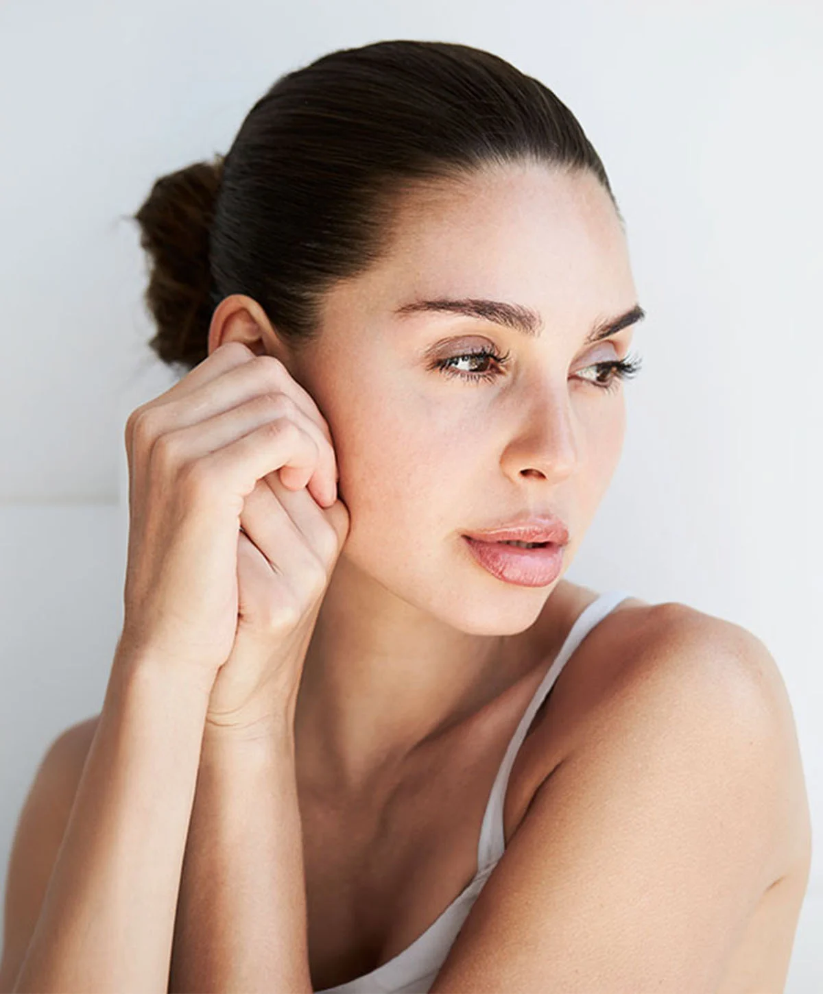 A woman with dark hair pulled back into a bun is looking away from the camera in profile. She is wearing a white tank top and gently touching the skin near her ear and jawline with both hands. - Deep Plane Facelift Recovery in Houston, TX