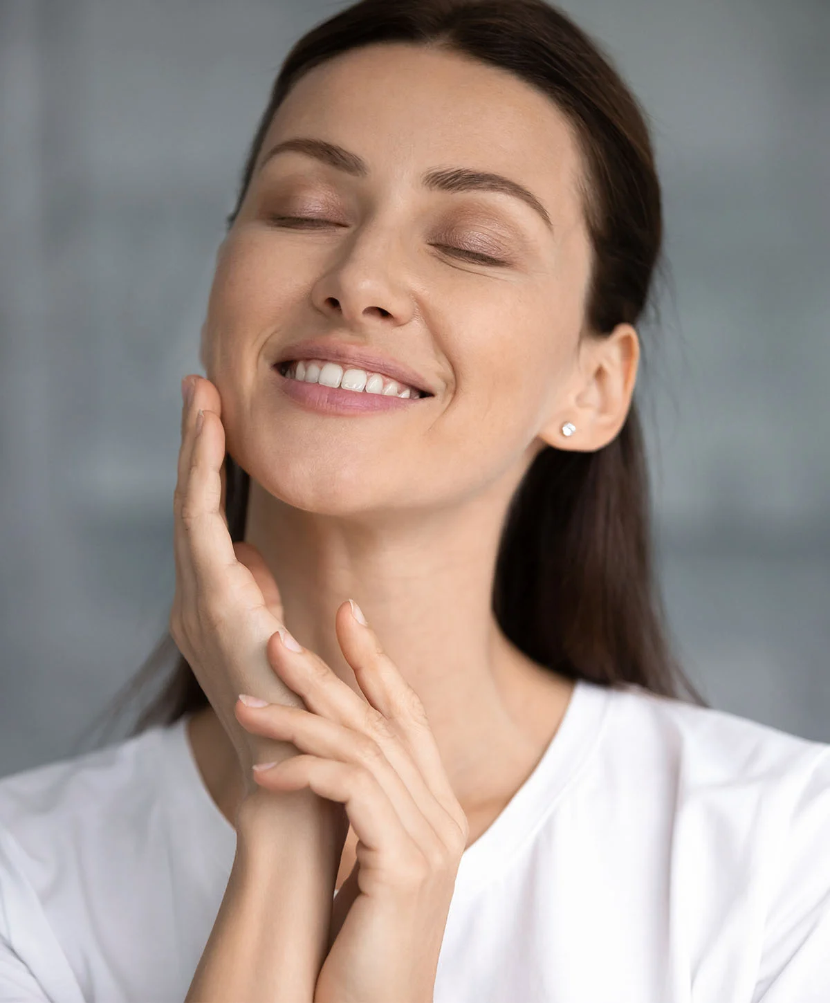 A young woman with long, dark hair looks directly at the camera with her eyes closed, gently touching her cheeks with both hands and smiling. She is wearing a white t-shirt, and the background is blurred, showing a light-colored, possibly tiled, surface. - Deep Plane Necklift Recovery in Houston, TX