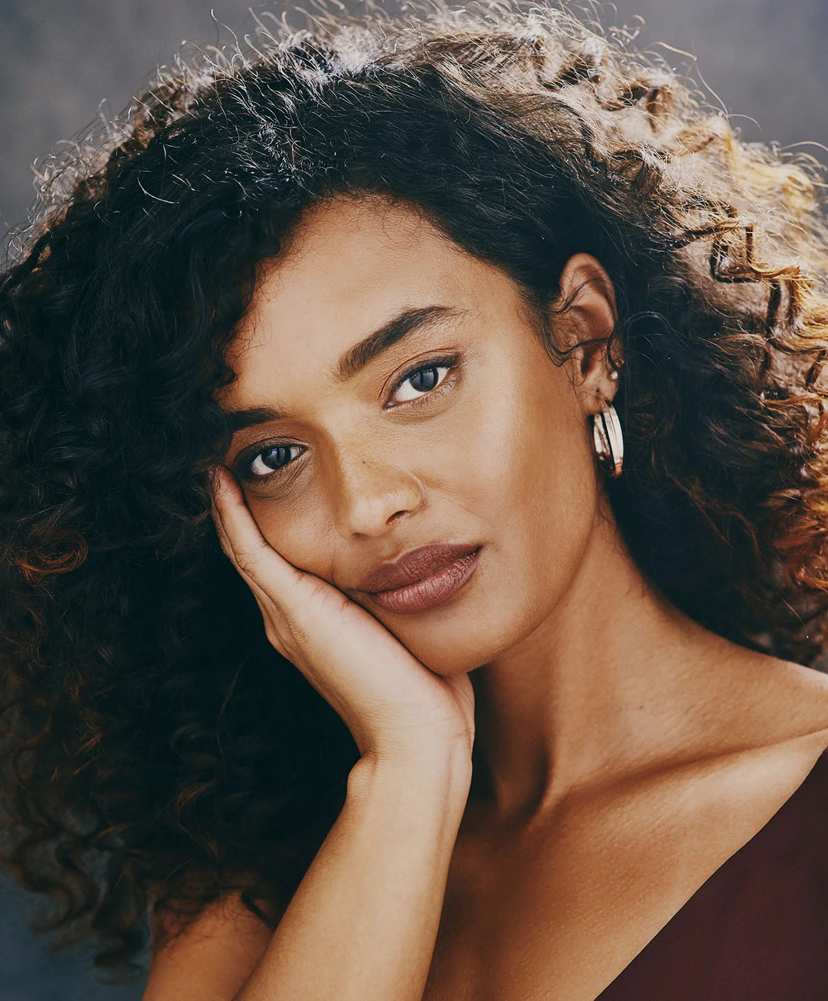 A stunning portrait of a woman with long, dark curly hair and warm skin looking directly at the camera. She is resting her chin on her hand and wearing a dark red top and a silver hoop earring. - Dermal Fillers in Houston, TX
