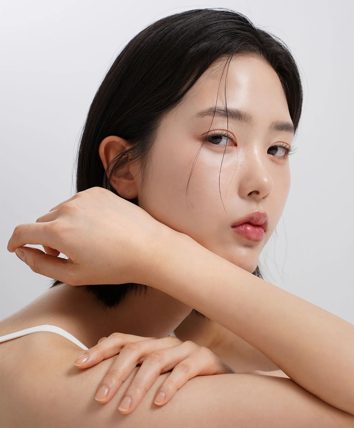 A portrait of a young woman with short, dark hair, wearing a white tank top, looking directly at the camera with a subtle gaze. She is resting her chin on one hand, and her other hand is touching her shoulder, against a white background. - Ethnic Rhinoplasty in Houston, TX