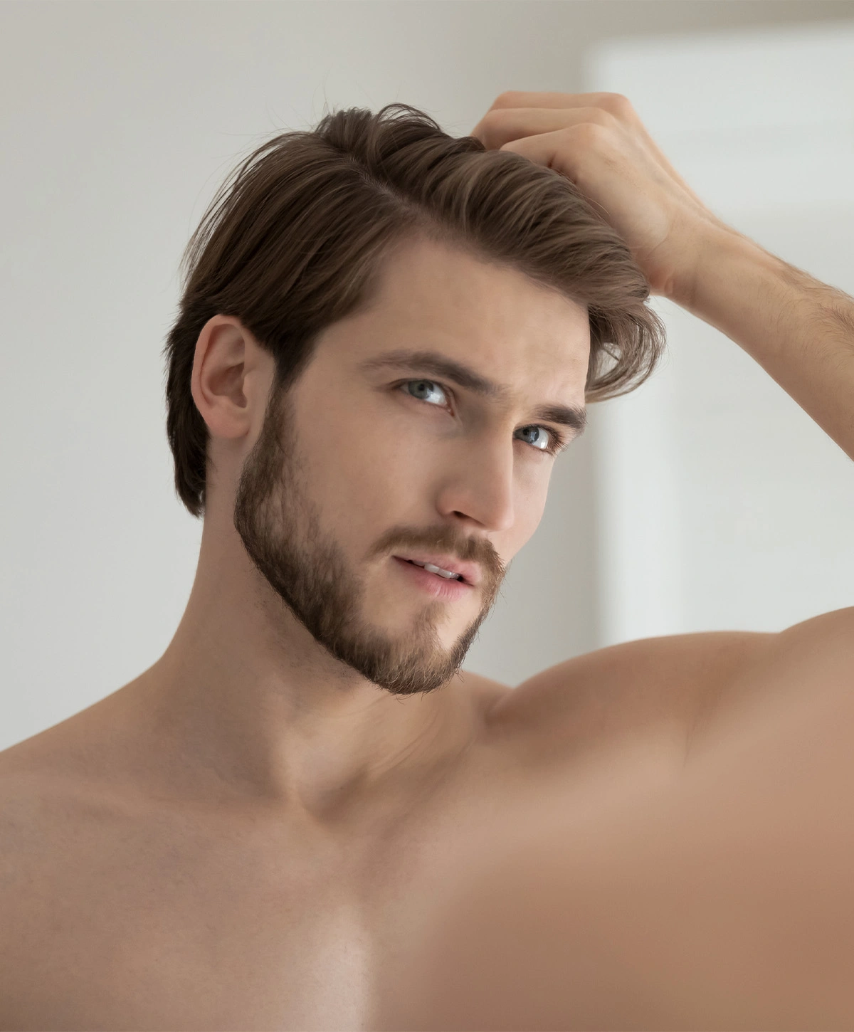 A man with light brown hair and a beard is shirtless and looking at his reflection while touching his hair. - Fue in Houston, TX