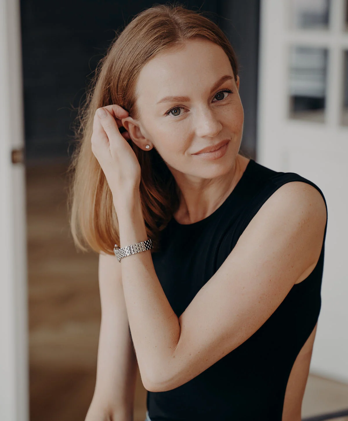 A beautiful woman with long, reddish-blonde hair is smiling and looking directly at the camera. She is wearing a black sleeveless top, a silver watch, and a small diamond earring, with one hand touching her hair near her ear. The background is an indoor setting with soft lighting. - Fut Hair Transplant in Houston, TX