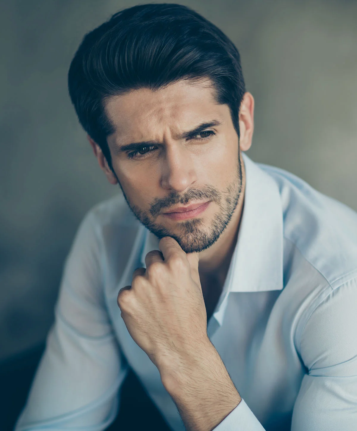 A close-up portrait of a man with dark hair and a short beard, wearing a light blue collared shirt. He has a serious expression and is resting his chin on his hand in a thoughtful pose. - Fut Hair Transplant Recovery in Houston, TX