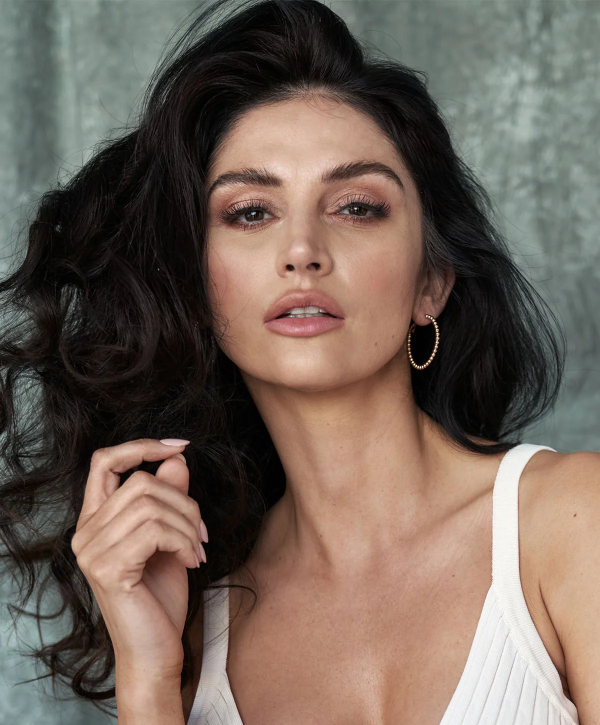 Close-up portrait of a light-skinned woman with long dark brown hair. She wears a cream-colored ribbed v-neck crop top, gold hoop earrings, and a gold chain bracelet. Her gaze is slightly downward with a direct expression. The background is a muted gray-green textured wall with soft lighting highlighting her features. - Lip Lift in Houston, TX