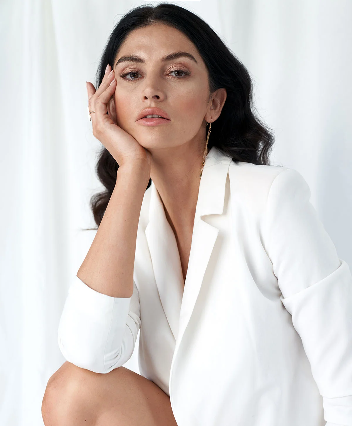A glamorous woman with long, dark, wavy hair is wearing a white blazer and posing against a white draped background. She is leaning her head on one hand and looking directly at the camera. - Liquid Facelift in Houston, TX