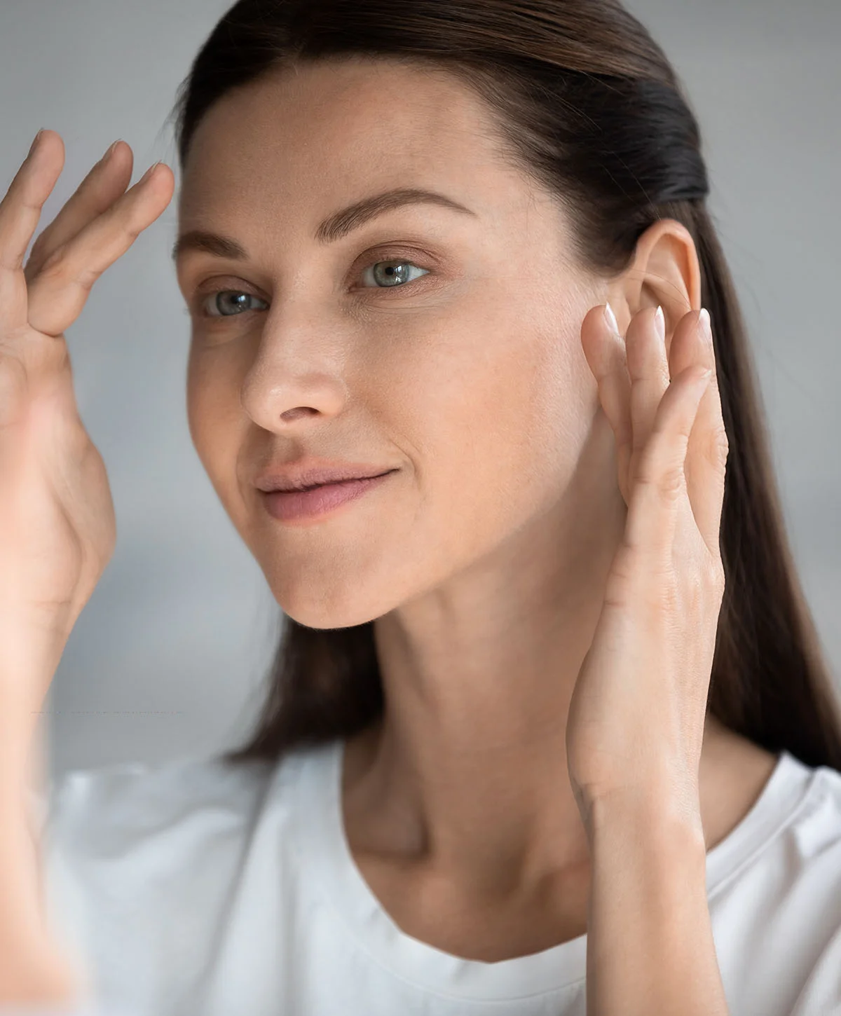 A woman with brown hair and light eyes is looking into a mirror, gently touching the sides of her face with her hands, possibly examining her skin. - Lower Blepharoplasty in Houston, TX