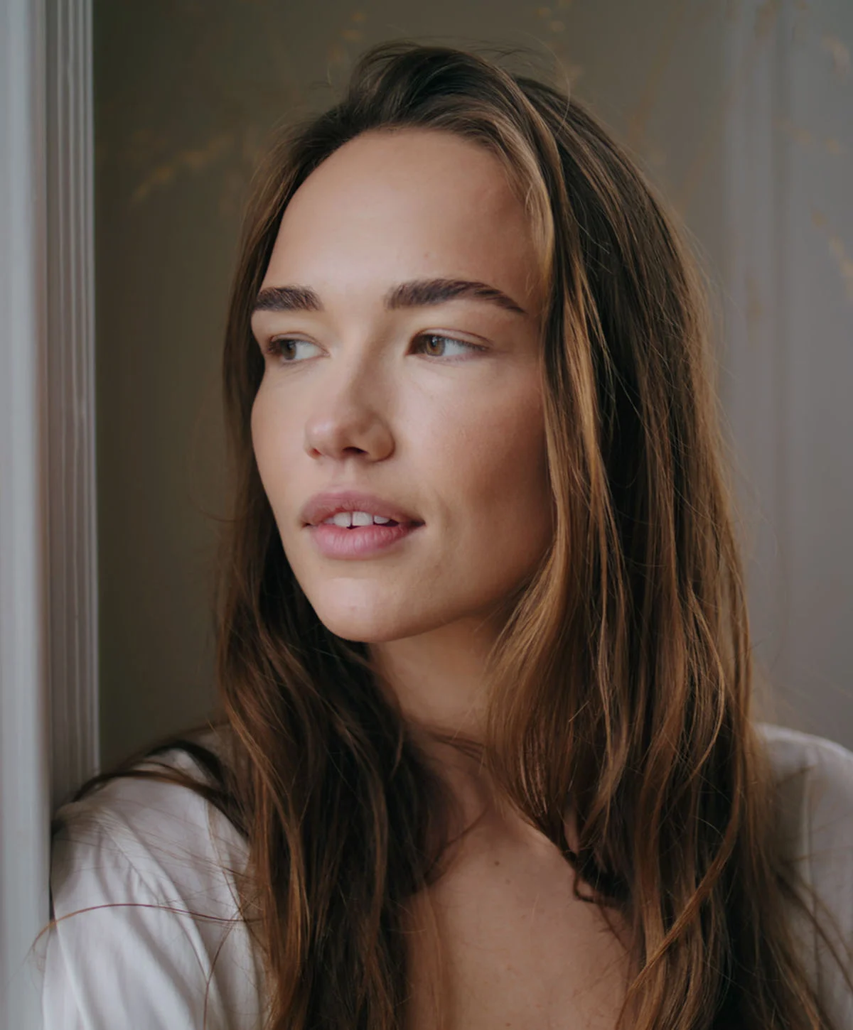 A natural portrait of a woman with long, flowing brown hair, wearing a white shirt, looking off to the side with a pensive expression. Soft, natural light falls on her face. - Lower Blepharoplasty Recovery in Houston, TX