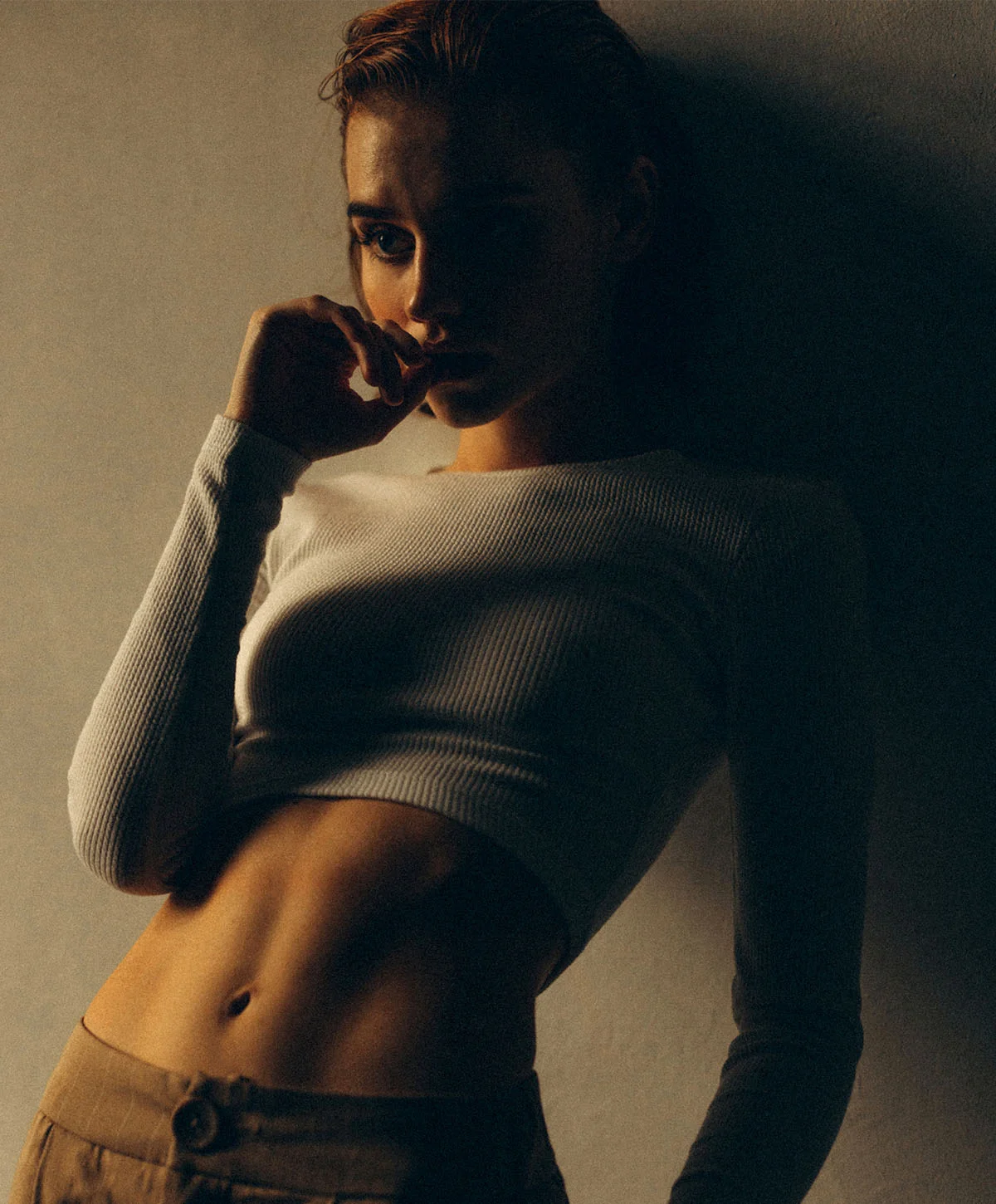 A moody, dramatic, low-angle shot of a woman with a fit physique leaning against a wall. She is wearing a white long-sleeve crop top and tan pants, with one hand near her mouth. The lighting is dark and contrasting. - Lympatic Drainage in Houston, TX