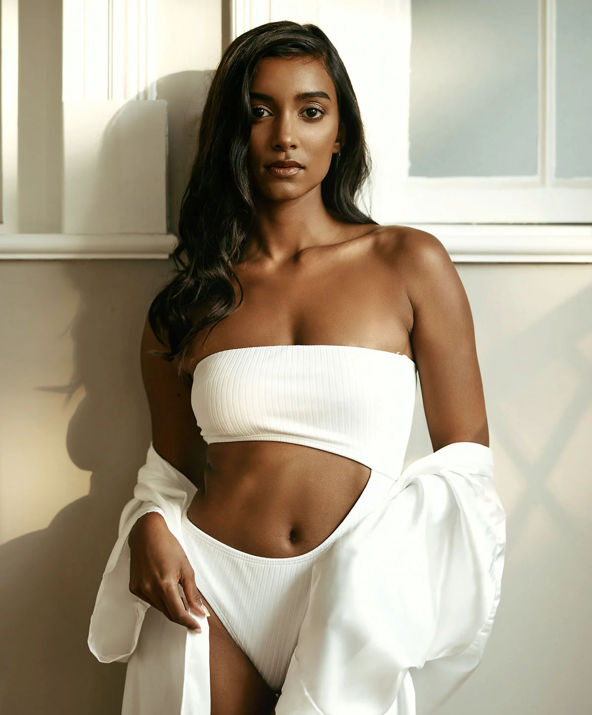 A tan-complexioned woman with long, dark brown hair stands in a softly lit room wearing a white two-piece swimsuit with a bandeau top and cutout bottom. A flowing white robe drapes over her shoulders and arms. The background includes light-colored arches and serene décor, creating a calm, luxurious atmosphere. Soft lighting highlights her figure. - Lympatic Drainage Recovery in Houston, TX