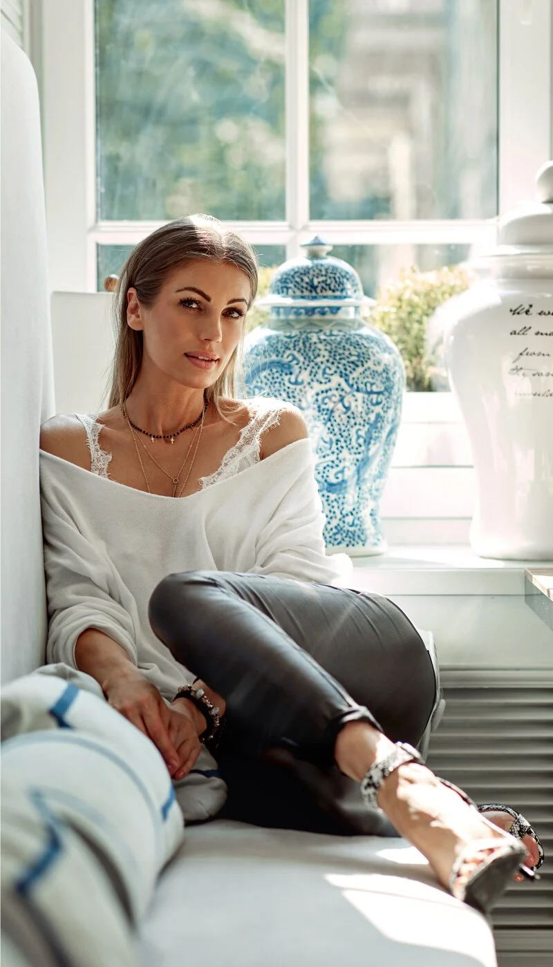 A fashionable woman is relaxing indoors on a window seat, wearing a white off-the-shoulder sweater over a lacy camisole and black leather-look trousers. She is looking directly at the camera, with a blue and white patterned vase and a large white vase on the windowsill behind her.