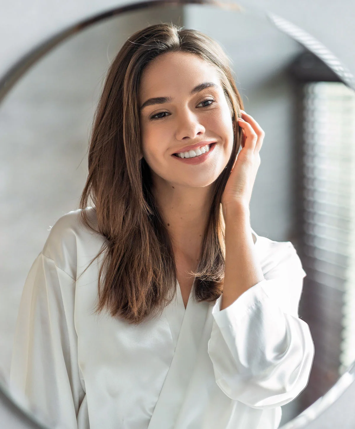 A smiling woman with long brown hair is reflected in a circular mirror, wearing a white silk robe and touching her hair near her ear. - Post Operative Care Kits in Houston, TX