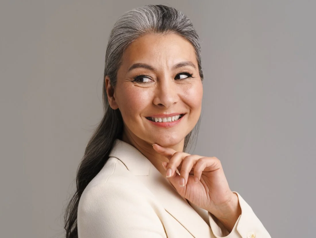 A middle-aged woman with long, salt-and-pepper hair, partially pulled back, is smiling and looking over her shoulder. She is wearing a light cream or white blazer and resting her chin on her hand against a plain gray background. - Surgical Procedures in Houston, TX