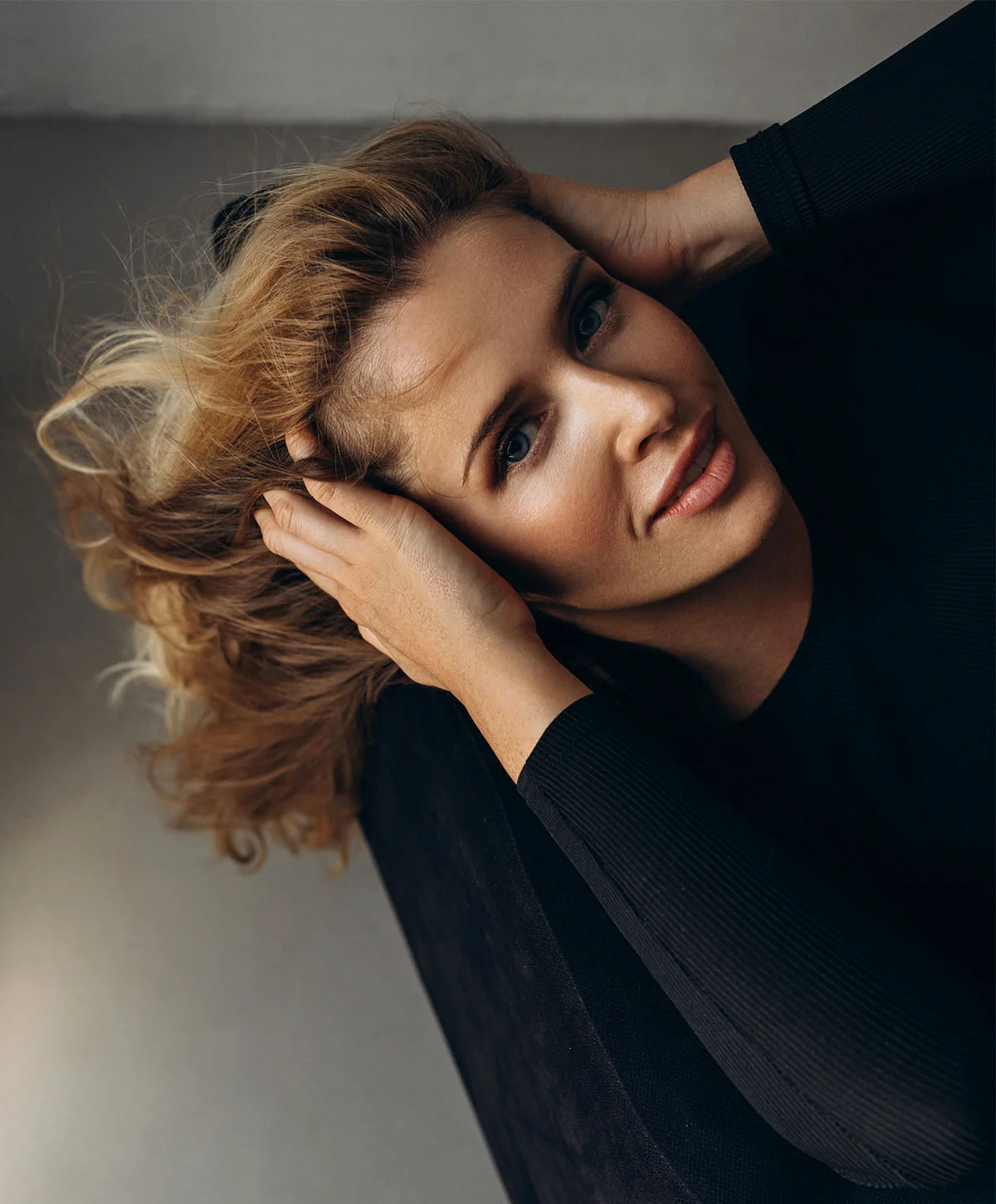 A portrait of a blonde woman with blue eyes, looking up at the camera while lying down or leaning back. She is wearing a black ribbed shirt and has both hands resting near her head. - Upper Blepharoplasty in Houston, TX