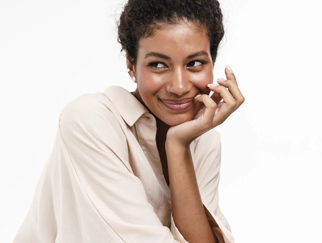 A close-up portrait of a woman with dark, curly hair pulled back, wearing a light beige collared shirt. She is resting her chin on her hand and looking off to the side with a bright, playful smile against a white background. - Wellness in Houston, TX