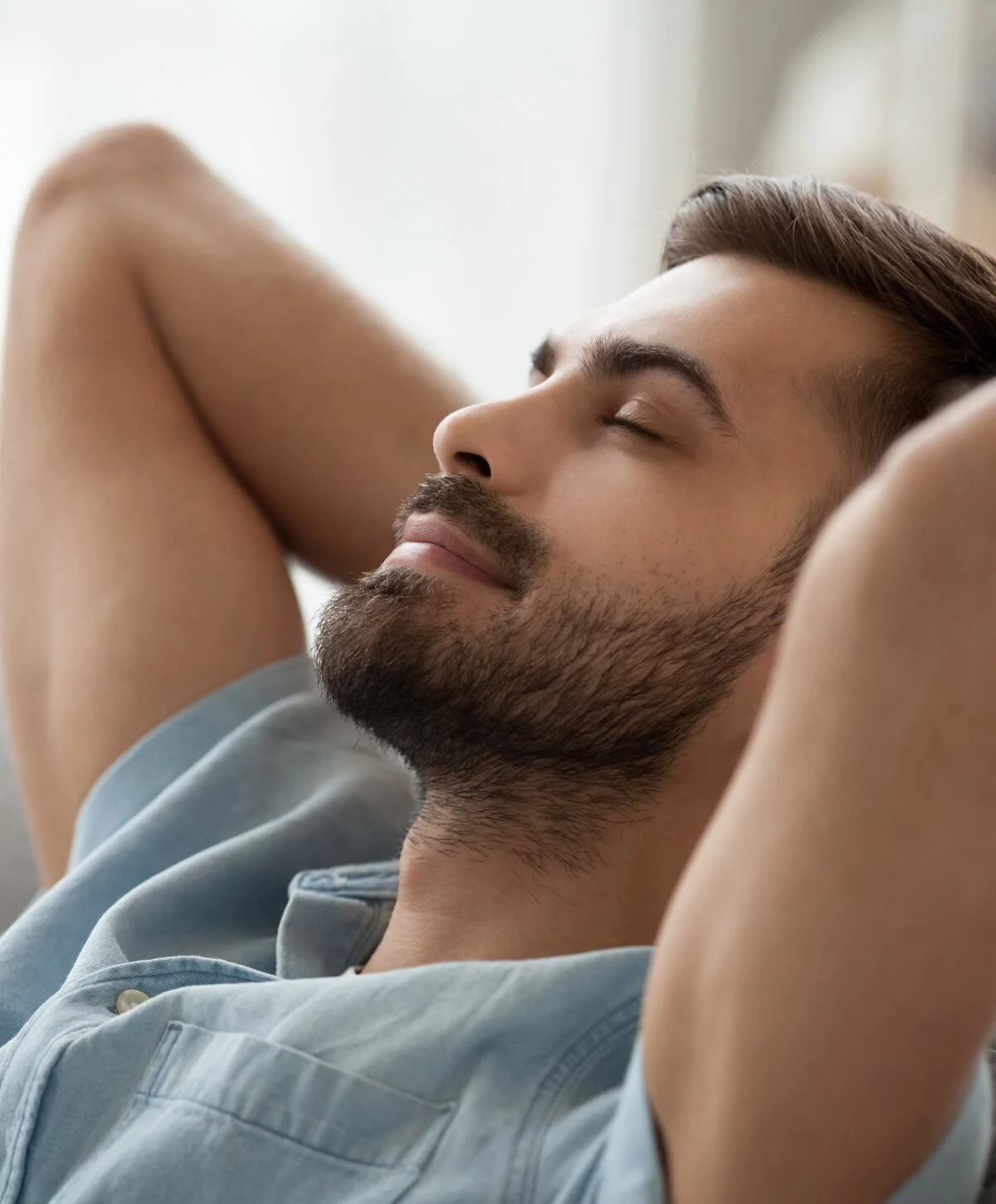 A close-up of a man with a beard and short hair, wearing a light blue shirt, relaxing with his eyes closed and his hands behind his head. He looks peaceful and calm. - Wellness in Houston, TX